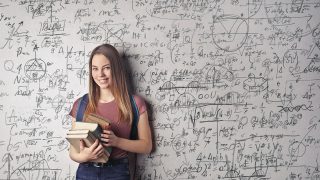 膨大な計算式などが書かれた白壁を背にハードカバーの本を数冊持って微笑む女性が立っている写真